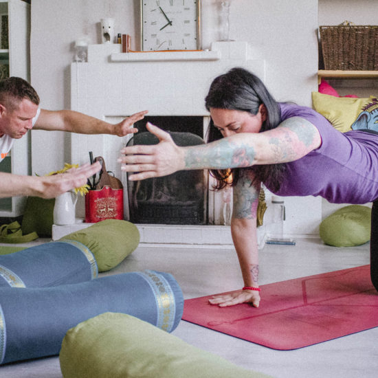 Two people practising side by side on a pink mat in a front room, arms reaching forward